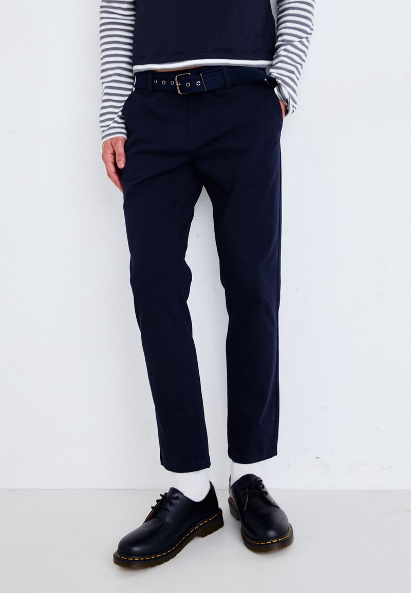 Marina bomullbyxor med slim fit, tillsammans med ett svart bälte med cirkulära hål. Bärs med svarta skor och vita strumpor. Randig långärmad topp.