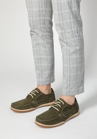 Olivgrüne Wildleder-Schuhe mit beigen Schnürsenkeln, Gummisohle und dekorativer Steppnaht. Graue Karohosen ergänzen das lässige Schuhdesign.