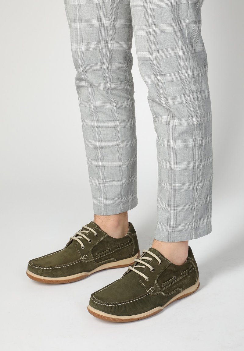 Olivgrüne Wildleder-Schuhe mit beigen Schnürsenkeln, Gummisohle und dekorativer Steppnaht. Graue Karohosen ergänzen das lässige Schuhdesign.