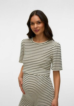 Gravid kvinde med langt mørkt hår iført en sort-hvid stribet kortærmet top og matchende nederdel, der forsigtigt holder sin mave.