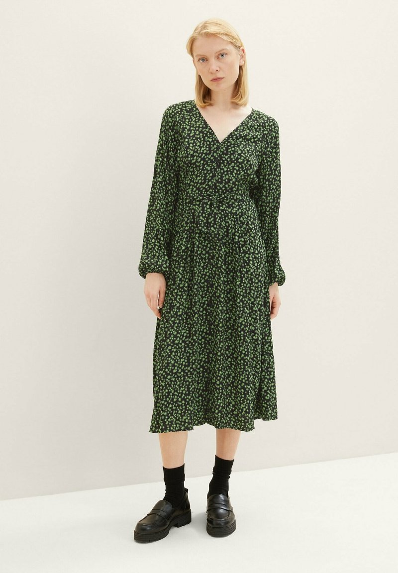 Kleid mit tiefem V-Ausschnitt, langen Ärmeln und taillierter Taille, aus dunkelgrünem Stoff mit einem kleinen Blumenmuster, knielang.