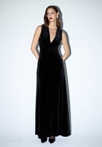 Lang zwart fluwelen jurk met een diep V-hals, mouwloos ontwerp en zijzakken. Vloeiende silhouet en subtiele glans, gedragen met zwarte hakken.