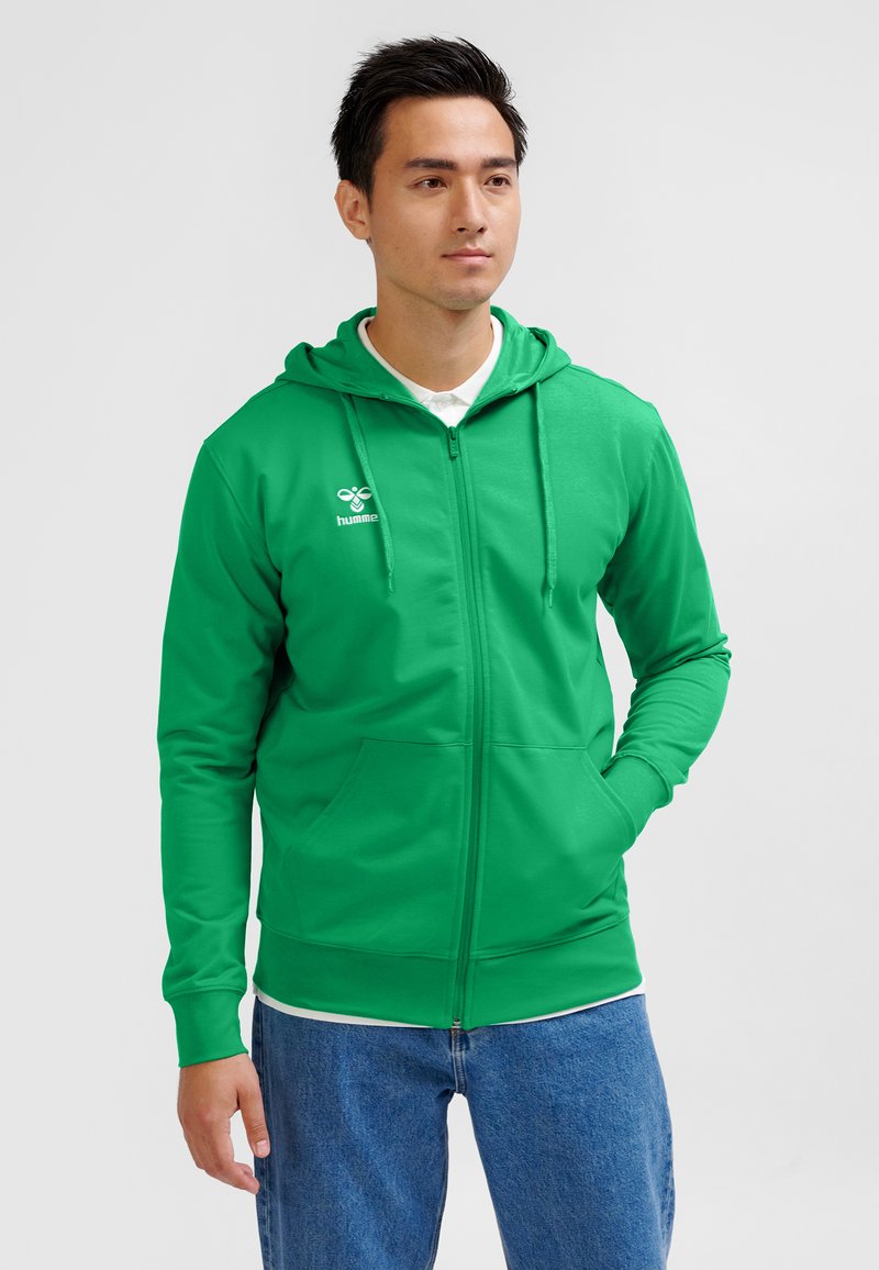 Grøn hættetrøje med lynlås, frontlomme, snore i hætten og hvidt logo på brystet; båret over en hvid skjorte og blå jeans.