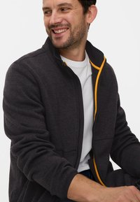 Dunkelgraue Reißverschlussjacke aus strukturiertem Stoff, mit einem kontrastierenden gelben Reißverschluss, Stehkragen und seitlichen Taschen.