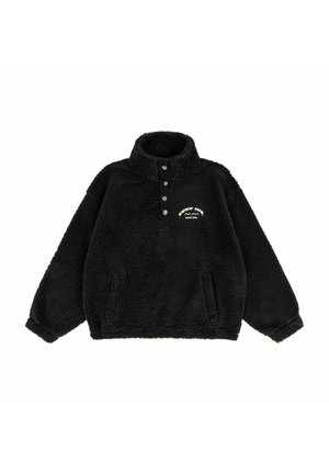 Kobieta w bordowej czapce i czarnej kurtce z logo "Surf Inn" uśmiecha się, a jej włosy powiewają na tle jasnego, zmierzchowego nieba.