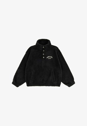 Kobieta w bordowej czapce i czarnej kurtce z logo "Surf Inn" uśmiecha się, a jej włosy powiewają na tle jasnego, zmierzchowego nieba.