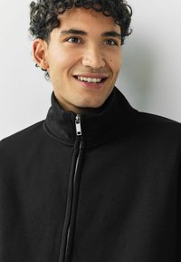 Junger Mann mit lockigem Haar, der lächelt und eine schwarze Zipfeljacke trägt, vor einem schlichten, hellen Hintergrund.