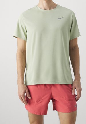 Mann trägt ein hellgrünes Nike-Sportshirt mit kurzen Ärmeln und korallenrote Sportshorts vor einem einfarbigen Hintergrund.