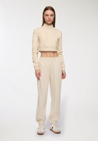 Giacca cropped cremosa con cerniera e colletto alto, dotata di tasca frontale, abbinata a pantaloni jogger con vita elastica e scarpe da ginnastica beige.
