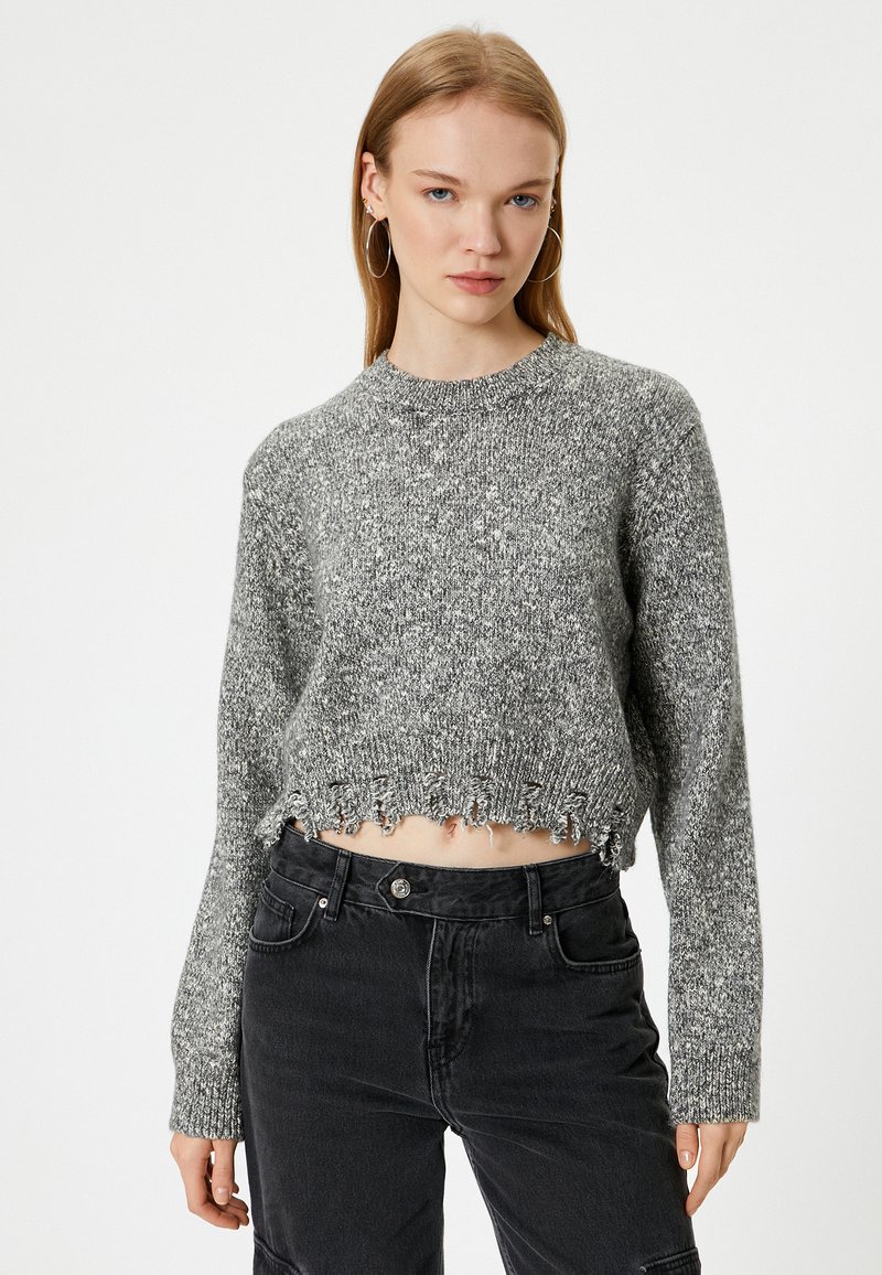 Korte trui in grijs breiwerk met een textuurpatroon, voorzien van een ronde halslijn en een rafelige onderkant. Gecombineerd met zwarte high-waisted jeans.