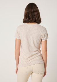 Ženska s temnimi ramenskimi lasmi, oblečena v svetlo bež majico s kratkimi rokavi in kremasto bele hlače, stoji obrnjena stran na enostavni podlagi.