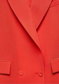 Blazer rouge à double boutonnière avec revers crantés et deux poches à rabat à l'avant, confectionné dans un tissu à la texture lisse.