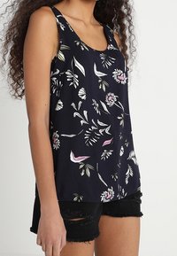 Granatowy top na ramiączkach z kwiatowym wzorem w bieli, różu i zieleni. Lekki materiał, okrągły dekolt, w połączeniu z czarnymi szortami z przetarciami.