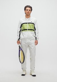 Νεαρός άνδρας με λευκή και πράσινη φόρμα που κρατά ρακέτα τένις, στέκεται μπροστά σε απλό λευκό φόντο.