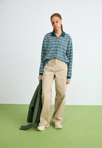Polo a righe, maniche lunghe, in teal e blu navy, abbinato a pantaloni larghi beige. Tiene in mano un giubbotto trapuntato verde. Calzature di colore chiaro.