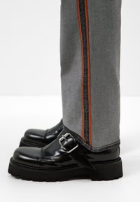 Zapatos gruesos de charol negro con hebilla plateada, combinados con jeans grises que tienen rayas laterales naranjas y negras.