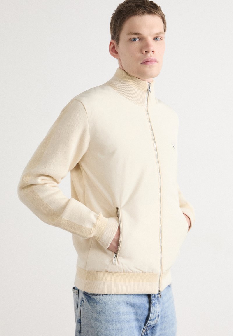 Beige zippered jacka med hög krage, ribbad mudd och sidofickor. Mjuk material med en figursydd design, kompletterad av ljus färg.