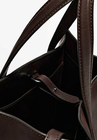 Bruine leren totebag met een gladde textuur, twee lange handvatten en zichtbare stiksels. Het interieur bevat vakken en een sluitband.