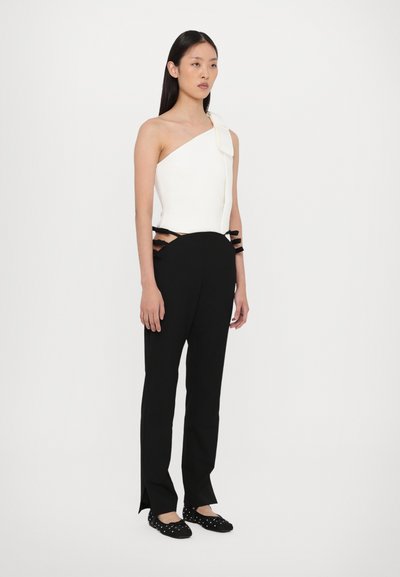 Witte one-shoulder top met een strikdetail, gecombineerd met zwarte getailleerde broek met uitsparingen en zwarte linten in de taille. Zwarte platte schoenen.
