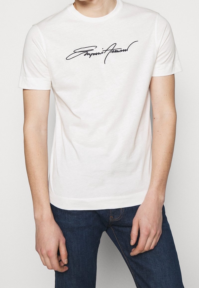 Vit bomullströja med en svart kursiv logotyp på framsidan. Korta ärmar, rundad hals och standard passform.