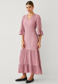 Rosekleurige maxi-jurk met een V-hals, driekwart fladdermouwen, textuurstof en een gelaagde zoom. Gepaard met zwarte flat shoes.
