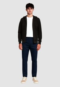 Felpa nera con zip, t-shirt bianca, pantaloni blu navy e sneakers beige. L'outfit presenta una vestibilità comoda, una texture liscia e un design semplice.