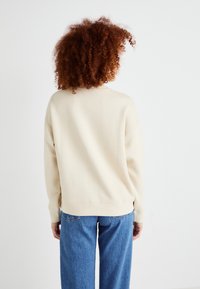 Beige Sweatshirt mit lockerer Passform, gerippten Bündchen und abgerundetem Saum, kombiniert mit hellblauen weiten Jeans. Hintergrund ist rein weiß.