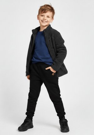 Uśmiechnięty chłopiec stoi z rękami w kieszeniach, ubrany w ciemną bluzę polarową, niebieską koszulę, czarne spodnie i czarne trampki na jednolitym tle.