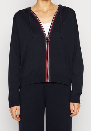 Personne portant un sweat à capuche zippé bleu marine avec des bordures rouge-blanc-bleu et un pantalon bleu marine assorti, debout devant un fond uni.
