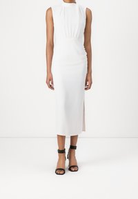 Robe blanche sans manches avec un col montant et une taille froncée, présentant une fente latérale et une texture lisse. Assortie à des talons noirs à bride à la cheville.