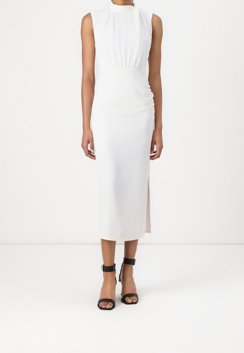Robe blanche sans manches avec un col montant et une taille froncée, présentant une fente latérale et une texture lisse. Assortie à des talons noirs à bride à la cheville.