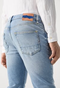 Lyseblå denimjeans med et brunt lærbelte bak, baklommer og fargerike sømdetaljer.