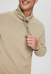 Pull à col montant beige en tricot avec un cordon de fermeture. Sa texture douce et ses poignets côtelés ajoutent du détail au design.