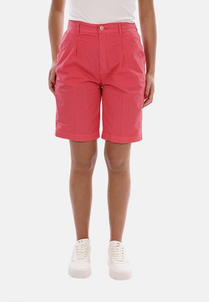 Femme portant un short corail taille haute arrivant aux genoux avec un bouton à l'avant et des baskets blanches, debout devant un fond uni.