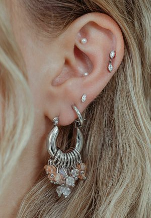 Silver hoop earring with dangling clear and peach-colored beads. Additional small silver studs are seen in the ear. Textured light hair is visible.