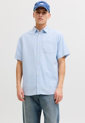 Joven con una camisa de manga corta azul claro abotonada, gorra azul y jeans holgados, de pie contra un fondo liso.