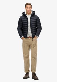 Chaqueta tipo plumas en color marino con costuras horizontales, diseño con capucha, llevada sobre un suéter gris claro, combinada con chinos caqui y botas marrones.