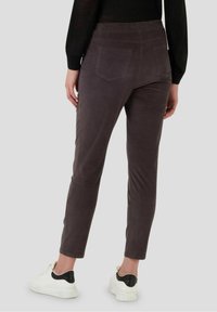 Žena v tmavě šedých úzkých kalhotách s zadními kapsami a bílých teniskách, stojící na jednobarevném světlém pozadí.