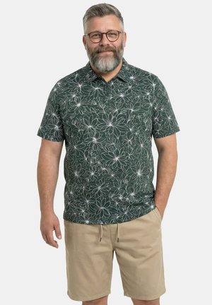 Mann mittleren Alters mit Brille und Bart, trägt ein dunkelgrünes kurzärmeliges Hemd mit Blumenmuster und beige Shorts, steht mit einer Hand in der Tasche.