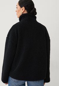 Zwarte fleece jack met hoge kraag, oversized pasvorm, textuurstof en verlaagde schouders. Draagt met lichtblauwe denim jeans.