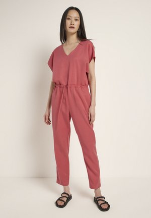 Pink jumpsuit with a V-neck and short sleeves, tapered legs, and an elastic waistband. Worn with black strappy sandals. Smooth texture.