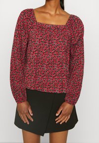 Blouse rouge à fleurs avec manches longues bouffantes, encolure carrée et texture froncée. À assortir avec une jupe noire à coupe asymétrique.