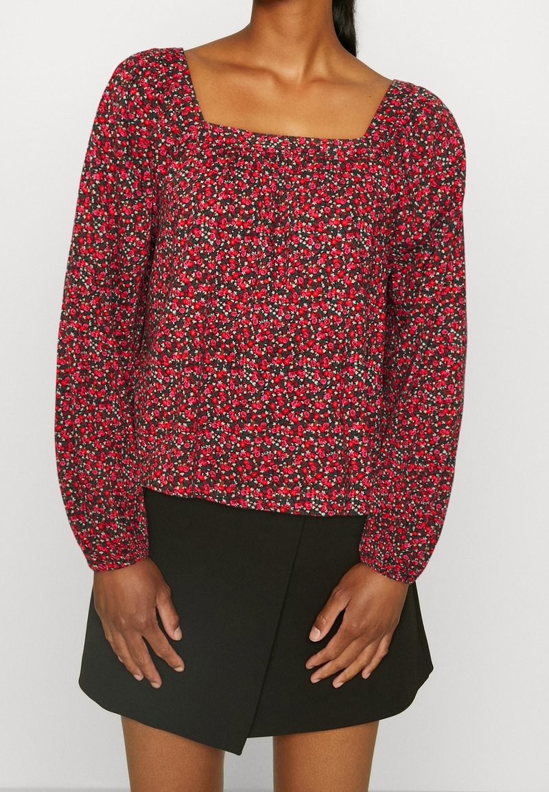 Blouse rouge à fleurs avec manches longues bouffantes, encolure carrée et texture froncée. À assortir avec une jupe noire à coupe asymétrique.