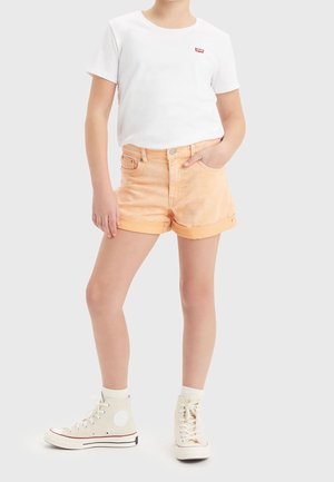 Weißes kurzärmeliges T-Shirt mit rotem Logo, kombiniert mit hellorangen Denim-Shorts. Beige High-Top-Sneaker mit weißen Akzenten runden den Look ab.