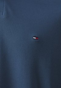 Blauwe polo met een gestructureerde stof; beschikt over een klein geborduurd logo in rood, wit en zwart op de borst.