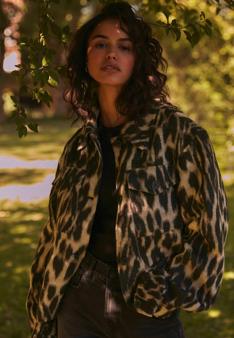 Leopardi-kuosi takki, jossa on teksturoitu, karvamainen pinta. Varustettu suurilla etutaskuilla ja tummilla napeilla, yhdistetty mustaan puseroon ja tummiin farkkuihin.