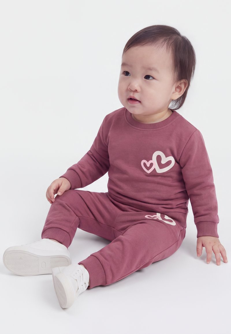 Tout-petit assis par terre portant un ensemble mauve à manches longues et pantalon avec des motifs de cœurs et des baskets blanches sur un fond blanc.