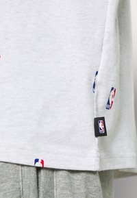 Szara koszulka z krótkim rękawem z małymi naszywkami z logo NBA w kolorach czerwonym, białym i niebieskim, charakteryzująca się miękką fakturą i prostym dołem.