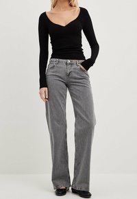 Schwarzes, geripptes Langarmoberteil mit V-Ausschnitt, kombiniert mit grauen, hoch geschnittenen weit geschnittenen Jeans, die über einen klassischen Knopf- und Reißverschluss verfügen.