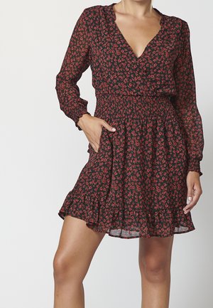 Sort langærmet kjole med rødt blomsterprint, smock-talje, V-halsudskæring, flæsekant, båret af en person med den ene hånd i lommen.